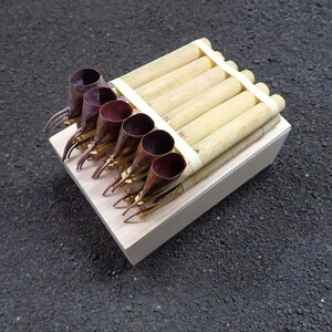 Può includere: Un set di sei utensili con manici in legno chiaro e punte coniche color rame scuro, disposti in fila e fissati con un elastico bianco. Gli utensili poggiano su una base rettangolare in legno chiaro.