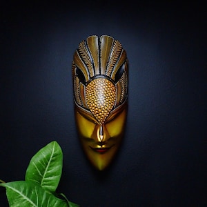 Op de afbeelding: Een gouden en zwart houten masker met ingewikkelde details en een vogelachtige snavel. Het masker heeft een menselijk gezicht met gesloten ogen en een kleine, rode mond.