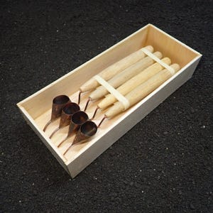 Peut inclure: Un ensemble de quatre outils tjanting avec des manches en bois clair et des réservoirs en cuivre, disposés dans une boîte en bois rectangulaire. Les outils sont maintenus ensemble par un élastique blanc, révélant leurs pointes métalliques.