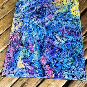 May include: Abstract art canvas with a vibrant mix of colors. The artwork features swirling patterns in shades of blue, purple, pink, and yellow, with black accents. The painting is displayed on a wooden surface.