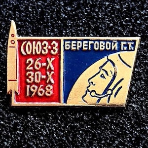 May include: A vintage pin with a rocket and astronaut design. The pin features text in Russian, including the date 1968. The pin has red, blue, and gold colors. The pin is rectangular with a pointed top.