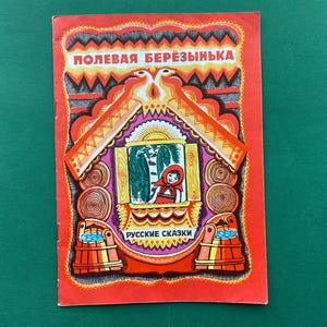 Pode incluir: Livro infantil vintage com capa vermelha. A capa apresenta o título "ПОЛЕВАЯ БЕРЁЗИНЬКА" e as palavras "РУССКИЕ СКАЗКИ". A ilustração mostra uma menina de capuz vermelho, uma janela e elementos decorativos.