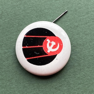 Puede incluir: Un botón redondo blanco con un diseño negro y rojo. El diseño incluye un círculo negro con tres líneas rojas y un emblema rojo con un martillo y una hoz blancos. Se adjunta un alfiler plateado.