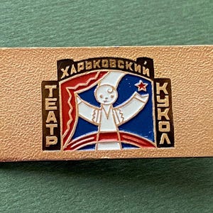 Puede incluir: Un pin rectangular con un fondo texturizado de color dorado. El pin presenta un diseño central con una figura de marioneta en blanco, rojo y azul, con texto en escritura cirílica. La palabra "TEATP" está verticalmente en el lado izquierdo, y "KUKOA" en el derecho.