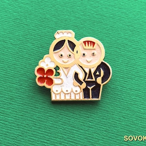 以下が含まれることがあります： 金色の金属製エナメルピン。白いドレスを着て赤と白の花束を持った人物と、王冠をかぶってダークスーツを着た人物の様式化されたイラストが描かれています。どちらも肌の色は明るく、黒髪です。