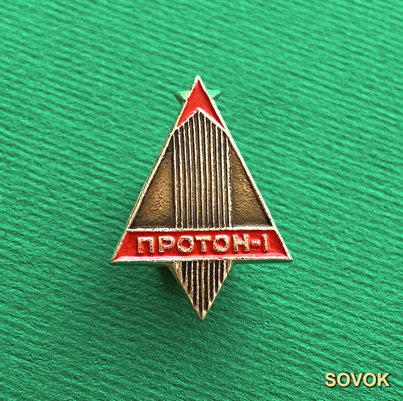 May include: A triangular metal pin featuring a gold-toned textured surface with vertical lines. The design includes a red enamel section at the top and a horizontal red enamel bar across the center with the Cyrillic text PROTON-1 in gold letters.