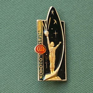 May include: A vintage gold-toned pin featuring a cosmonaut reaching for a globe, set against a black backdrop with stars and a stylized rocket. The pin includes vertical text and a red and orange circular emblem.