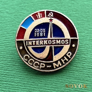 May include: A circular metal pin featuring a blue center with a rocket graphic, the text 22.03 1981, and the word INTERKOSMOS. The border includes red, blue, and dark sections with the symbols of a hammer and sickle and the text CCCP-MHP in gold lettering.