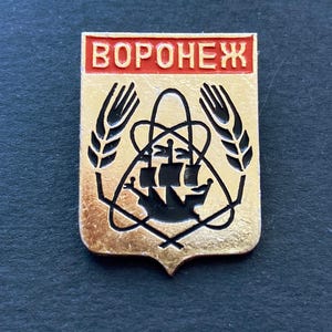 May include: A gold-colored pin with a red rectangular top that reads "VORONEZH". The pin features a black ship, wheat stalks, and other geometric shapes. The pin is on a dark surface.