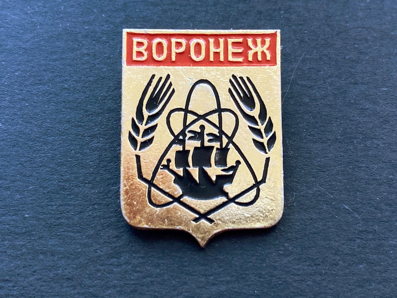 May include: A gold-colored pin with a red rectangular top that reads "VORONEZH". The pin features a black ship, wheat stalks, and other geometric shapes. The pin is on a dark surface.