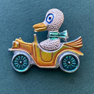 May include: A whimsical brooch featuring a duck driving a vintage car. The duck has a textured head, blue eyes, and a green scarf. The car is yellow and orange with teal wheels, creating a charming, vintage-inspired accessory.