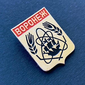May include: A vintage pin with a shield shape, featuring the word "Воронеж" in red letters at the top. The pin has a gold background with black graphic elements, including wheat stalks and a stylized atom symbol. The pin is likely a collectible.