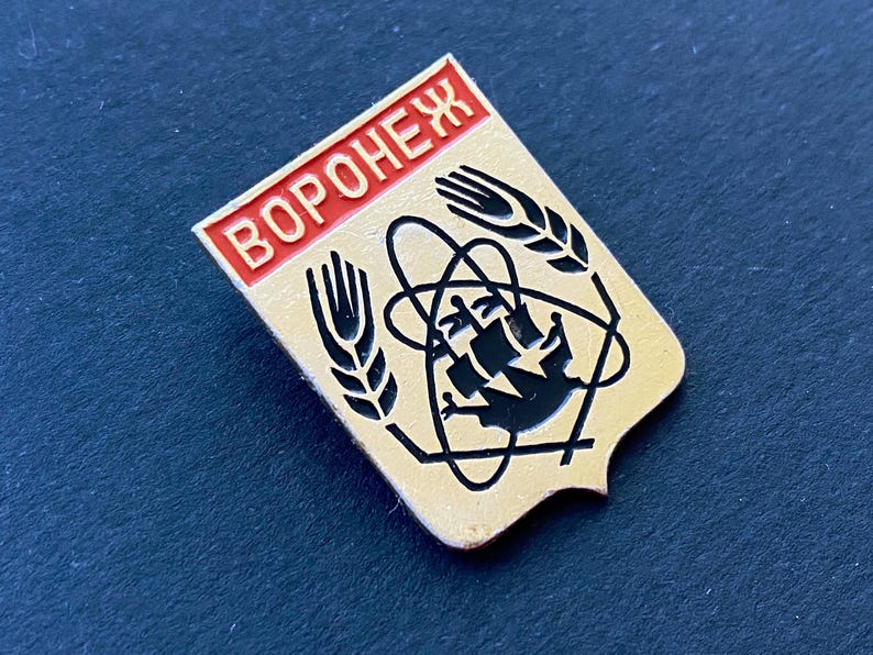 May include: A vintage pin with a shield shape, featuring the word "Воронеж" in red letters at the top. The pin has a gold background with black graphic elements, including wheat stalks and a stylized atom symbol. The pin is likely a collectible.