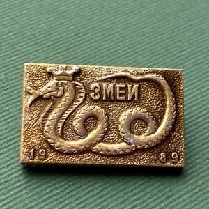 Puede incluir: Un pin rectangular dorado con un diseño detallado de serpiente coronada, la palabra cirílica "ЗМЕИ" y los números "19" y "89". El pin está sobre un fondo verde texturizado.