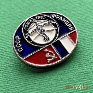 May include: An oval metal pin featuring a central circular emblem with a space station graphic and the year 1982. The design includes the text CCCP, SOYUZ, and FRANCIA, alongside a red, white, and blue flag motif and a silver hammer and sickle symbol.