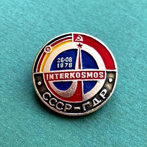 May include: A round, vintage Interkosmos pin with a red, blue, yellow, and black design. The pin features the text "INTERKOSMOS" and "26-08 1978". The pin also has the text "CCCP-ГДР" in a circular pattern.