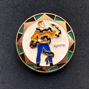 Könnte beinhalten: Runder, vintage Ansteckpin mit Zirkusmotiv. Der Pin zeigt einen Mann in einer gelben Jacke und blauen Hose, der eine Schlange hält. Der Pin hat einen weißen Hintergrund mit dem Wort "цирк" und ist von goldenen, grünen und schwarzen geometrischen Formen umgeben.
