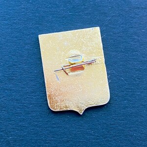 May include: A gold-colored pin with a shield shape and a pin back. The pin has a rectangular top and a pointed bottom. The pin back is a safety pin style with a red bar and a gold-colored clasp. The pin is on a dark blue surface.