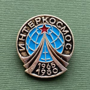 May include: Vintage Interkosmos pin with a blue globe design, a red star, and the years 1965 and 1980. The pin is circular, with a gold-colored metal finish and blue enamel details. The pin is likely a collectible item.