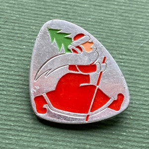 Può includere: Uno spillo natalizio argentato con un Babbo Natale stilizzato che porta un piccolo albero di Natale verde e una borsa rossa. Lo spillo ha la forma di una lacrima ed è di colore rosso, bianco e verde.