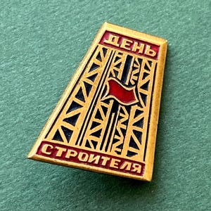 Puede incluir: Un pin trapezoidal de metal dorado con un diseño central de torre de celosía en negro y oro con una bandera roja. La banda roja superior muestra el texto cirílico DENE, y la banda roja inferior muestra el texto cirílico STROITELYA.
