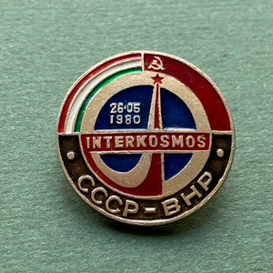 May include: A circular metal pin featuring a red, white, green, and blue enamel design. The center displays a rocket graphic, the text "INTERKOSMOS", "26-05 1980", and "CCCP-BHP". A small hammer and sickle symbol is positioned at the top edge.