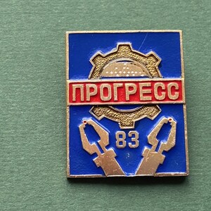 Puede incluir: Pin rectangular azul y dorado con la palabra rusa "PROGRESS" en letras rojas. El pin presenta un engranaje dorado, dos herramientas doradas y el número "83".