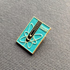 Peut inclure: Une &eacute;pingle rectangulaire en &eacute;mail turquoise avec une bordure dor&eacute;e. L'&eacute;pingle pr&eacute;sente un motif d'atome noir et or et les lettres cyrilliques "BAHX". L'&eacute;pingle est pos&eacute;e sur une surface grise textur&eacute;e.