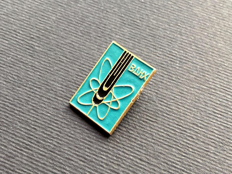 Peut inclure: Une &eacute;pingle rectangulaire en &eacute;mail turquoise avec une bordure dor&eacute;e. L'&eacute;pingle pr&eacute;sente un motif d'atome noir et or et les lettres cyrilliques "BAHX". L'&eacute;pingle est pos&eacute;e sur une surface grise textur&eacute;e.