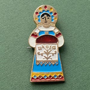 May include: A gold-toned pin featuring a woman in a traditional Russian folk costume. She is wearing a blue and white dress with a red and white floral pattern. Her head is covered with a red and yellow patterned headdress.