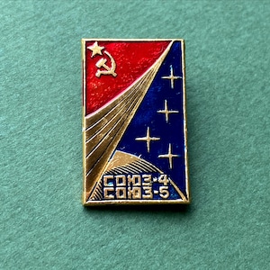 May include: Rectangular vintage pin with a red and blue design. The top left corner features a gold star, hammer, and sickle. The right side has four white stars on a blue background. Gold lines and text are also present.