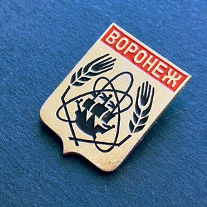 May include: Vintage pin with the word "VORONEZH" in red lettering. The pin features a gold background with black and white graphics, including wheat stalks and a stylized atom symbol. The pin is likely a collectible item.