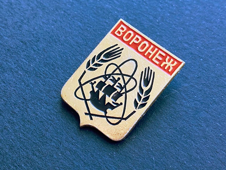 May include: Vintage pin with the word "VORONEZH" in red lettering. The pin features a gold background with black and white graphics, including wheat stalks and a stylized atom symbol. The pin is likely a collectible item.
