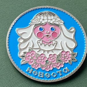 以下が含まれることがあります： 青と銀色の丸いピンバッジ。白いベールとピンクの頬をした花嫁が描かれています。花嫁はピンクの花に囲まれ、ロシア語で「Невеста」（花嫁）という言葉が青色で書かれています。