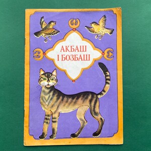 Pode incluir: Capa de livro vintage com uma ilustração de gato tigrado sobre fundo roxo. O texto "АКБАШ I БОЗБАШ" é exibido em uma moldura decorativa, com dois pássaros acima. A capa tem uma borda laranja.