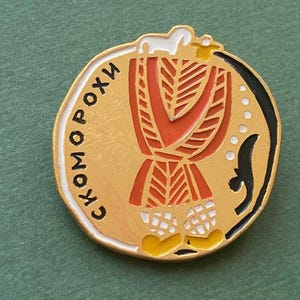 Pode incluir: Um alfinete dourado com um design vermelho e branco. O alfinete apresenta uma figura estilizada com uma cabeça branca e um corpo com um padrão vermelho e branco. O texto "СКОМО РОХИ" está escrito em preto no alfinete.