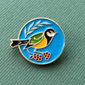 May include: A round, gold-edged pin featuring a colorful bird design. The bird has a yellow breast, green wings, and a black tail, set against a bright blue background. A sprig of leaves is above the bird, and red berries are below.
