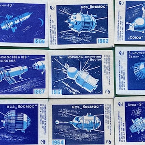 May include: A collection of nine vintage matchbook covers featuring illustrations of Soviet space exploration. The covers are blue and white with text in Russian and Cyrillic characters. The text includes the names of various space missions, such as "Luna-10", "Kosmos 186 and 188", "Soyuz", and "Luna-9".
