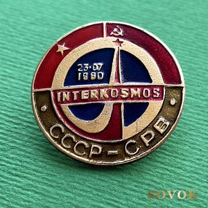 May include: A circular metal pin featuring a red, blue, and gold design. The center displays the text INTERKOSMOS, the date 23.07 1980, and a rocket icon. The bottom rim includes the Cyrillic text CCCP-CPB, with a hammer and sickle symbol at the top.