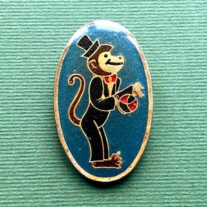 May include: An oval pin depicting a cartoon monkey in a black suit, top hat, and red bow tie. The monkey holds a ball with red and black stripes. The background is blue with a gold border.