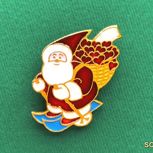 May include: A gold-toned metal enamel pin featuring Santa Claus skiing on bright blue skis. Santa wears a deep red suit and carries a yellow basket filled with numerous small red hearts on his back. The design includes white accents for the beard.