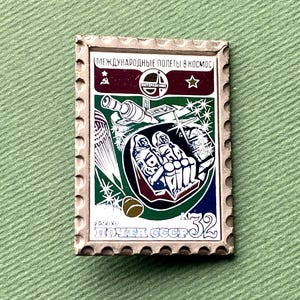 May include: Vintage Soviet-era postage stamp featuring a space exploration theme. The stamp has a rectangular shape with a decorative border. The design includes a spacecraft, astronauts, and text in Russian. The stamp is primarily green, red, white, and blue.