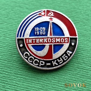May include: A circular metal pin featuring a red, white, and blue enamel design. It displays the text INTERKOSMOS, 18-09 1980, and CCCP-КУБА. The emblem includes a hammer and sickle symbol, a star, and a rocket graphic against a blue background.