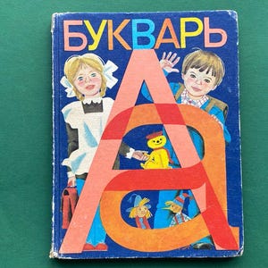 May include: Vintage children's book with a blue cover featuring the word "БУКВАРЬ" in yellow and red letters. The cover illustration includes two children, a large letter "A", and other cartoon characters. The book has a worn appearance.