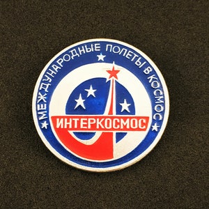 May include: A circular metal pin featuring a blue and white design with a red horizontal bar. The text "МЕЖДУНАРОДНЫЕ ПОЛЕТЫ В КОСМОС" is written in white along the top edge, and "ИНТЕРКОСМОС" is printed in white on the red bar. A red star sits above.