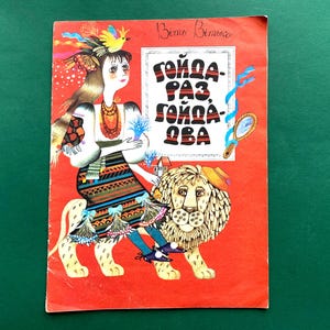 May include: Vintage children's book cover with a red background. The illustration features a woman with long hair, a bird, and a lion. The text on the cover is in a bold, stylized font. The book cover has a folk art style.