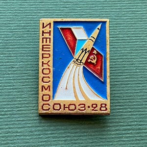 May include: A rectangular metal pin featuring a blue background with a white rocket launching upward. The design includes a red flag with a gold hammer and sickle, and the Cyrillic text "ШЕЙЕРУКОСМОС" vertically and "СОЮЗ-28" horizontally in gold.
