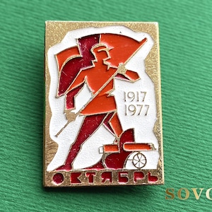 May include: A rectangular gold-tone metal pin featuring a red and orange enameled figure of a soldier holding a flag. The design includes the years 1917 and 1977, a small cannon, and the Cyrillic text ОКТЯБРЬ in red enamel at the bottom edge.