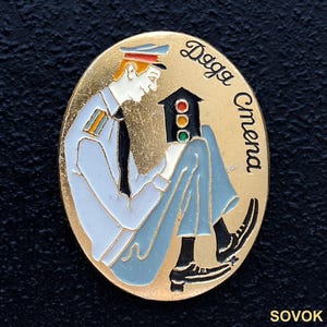 May include: Oval-shaped gold-tone pin featuring a uniformed figure holding a traffic light. The figure is in light blue with a black tie and cap. The pin has the text "Daga Cmena" in black and the word "SOVOK" at the bottom.