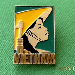 Op de afbeelding: Een rechthoekige metalen speld met een profielillustratie van een persoon die een gele kegelvormige hoed draagt tegen een donkergroene achtergrond. Links staat een goudkleurige structuur naast de tekst VIETNAM in gouden hoofdletters.